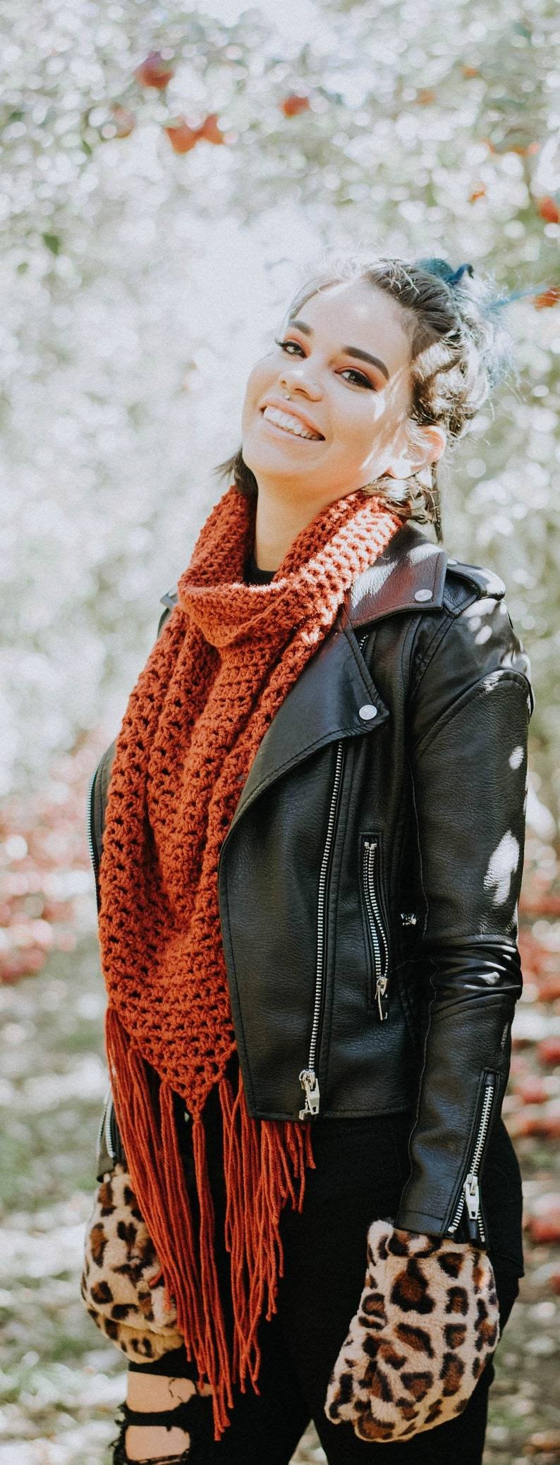 May include: A woman wearing a black leather jacket with a rust-colored crocheted scarf and leopard print mittens.
