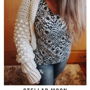 Op de afbeelding: Een close-up van een vrouw die een wit gebreid vest draagt met een textuur, bobbelsteekpatroon. Het vest heeft lange mouwen en is getailleerd. De vrouw draagt ook een zwart-wit geprint shirt en een blauwe jeans. De tekst "STELLAR MOON crochet pattern serendipity-as-always.com" is zichtbaar onderaan de afbeelding.