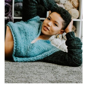 May include: A woman wearing a blue and black crochet hoodie. The hoodie has a V-neck and long sleeves. The woman is lying on a gray carpet. The text "HARMONY HOODIE" and "crochet pattern" is visible below the image. The text "serendipity-as-always.com" is visible below the text.