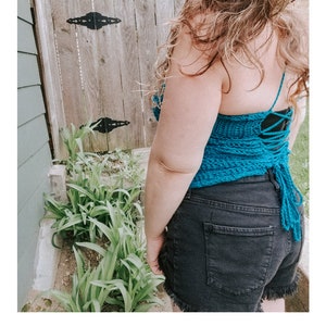 May include: A teal blue crochet halter top with a lace-up back. The top is worn by a person with long brown hair. The top is made with a shell stitch pattern.