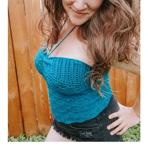 May include: A teal crochet halter top with a criss-cross strap design. The top is worn by a person with long brown hair. The text "BOMB SHELL HALTER" is at the bottom of the image. The text "crochet pattern" is below the first line of text. The text "serendipity-as-always.com" is below the second line of text.