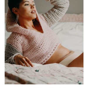 May include: A pink and gray crochet hoodie with a hood. The hoodie is cropped and has a v-neck. The model is wearing the hoodie on a bed with a floral patterned bedspread. The text "HARMONY HOODIE" and "crochet pattern" are visible below the image. The text "serendipity-as-always.com" is visible below the text.
