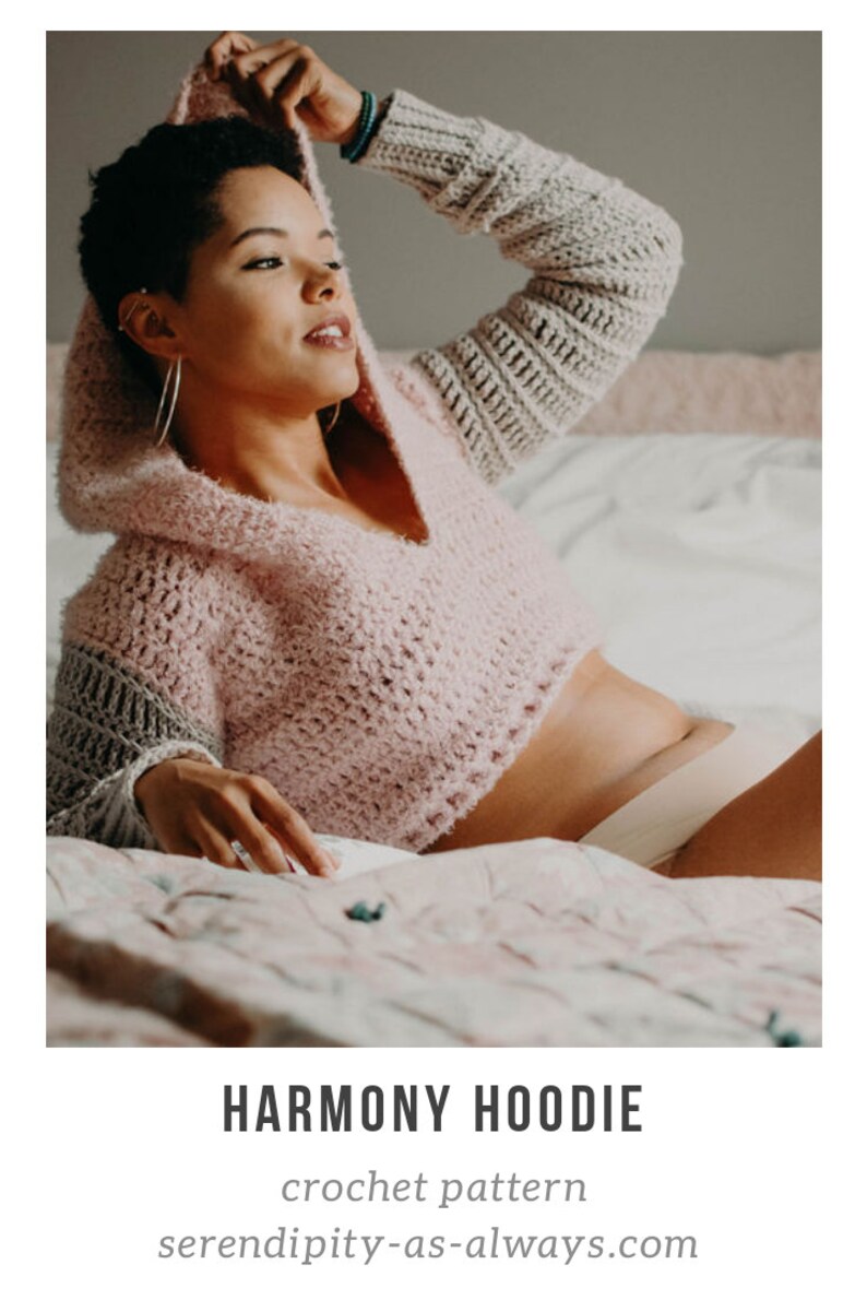 May include: A pink and gray crochet hoodie with a hood. The hoodie is cropped and has a v-neck. The model is wearing the hoodie on a bed with a floral patterned bedspread. The text "HARMONY HOODIE" and "crochet pattern" are visible below the image. The text "serendipity-as-always.com" is visible below the text.