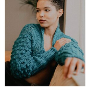 Op de afbeelding: Een tealblauwe gehaakte sjaal met een textuurpatroon. De sjaal wordt gedragen door een persoon met kort haar en is gestyled met een casual, ontspannen look. De tekst "STELLAR MOON SHRUG" en "crochet pattern" is zichtbaar onder de afbeelding. Het websiteadres "serendipity-as-always.com" is ook zichtbaar.