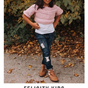 May include: A young person wearing a pink crochet top with short sleeves, blue jeans, and brown boots. The top is a cropped style and the jeans have distressed details. The person is standing in a wooded area with fallen leaves on the ground. The text "FELICITY KIDS crochet pattern" and "serendipity-as-always.com" is visible at the bottom of the image.