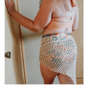 May include: A crocheted sarong in a beige color with a floral pattern. The sarong is tied around the waist and drapes down the legs. The text "SERENDIPITY SARONG" and "crochet pattern" is visible at the bottom of the image. The website address "serendipity-as-always.com" is also visible.