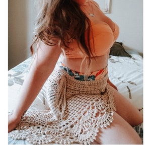 May include: A crocheted sarong in a beige color with a delicate lace pattern. The sarong is draped over a person's legs and hips. The text "SERENDIPITY SARONG" and "crochet pattern" is visible below the image. The website address "serendipity-as-always.com" is also visible.