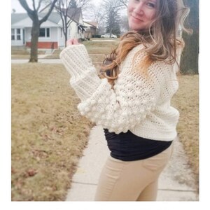 Op de afbeelding: Een witte gehaakte shrug met een textuurpatroon en pofmouwen. De shrug wordt gedragen door een persoon met lang bruin haar. De tekst "STELLAR MOON SHRUN crochet pattern serendipity-as-always.com" is zichtbaar onderaan de afbeelding.
