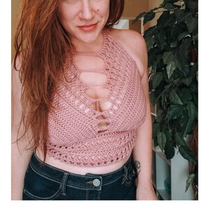 May include: A woman wearing a pink crochet halter top with a cutout design. The top is cropped and has a loose fit. The woman is also wearing blue jeans.