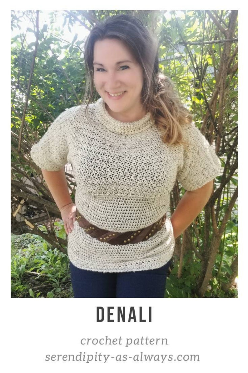 May include: A white crochet top with a brown leather belt with gold studs. The top has a round neckline and short sleeves. The model is standing in front of a green bush.