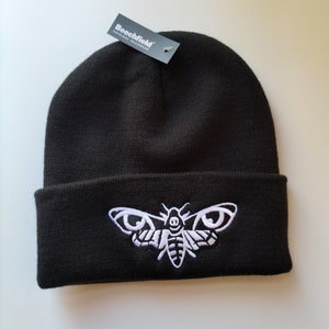May include: Black beanie with a white embroidered moth design. The beanie has a fold-over cuff and a tag that reads "Beechfield Original Headwear".