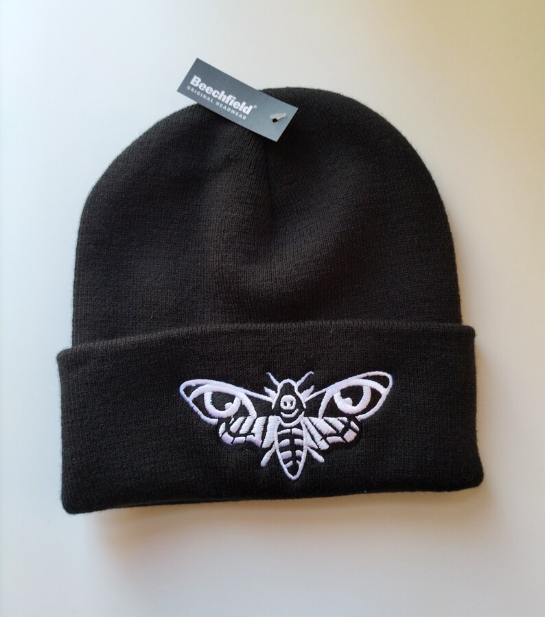 May include: Black beanie with a white embroidered moth design. The beanie has a fold-over cuff and a tag that reads "Beechfield Original Headwear".