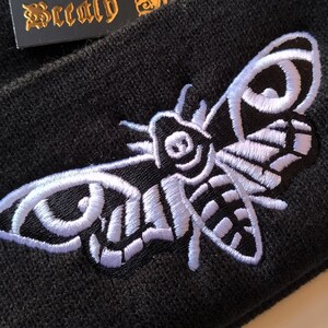 May include: Black beanie with a white embroidered moth design. The moth has a large wingspan and intricate details.