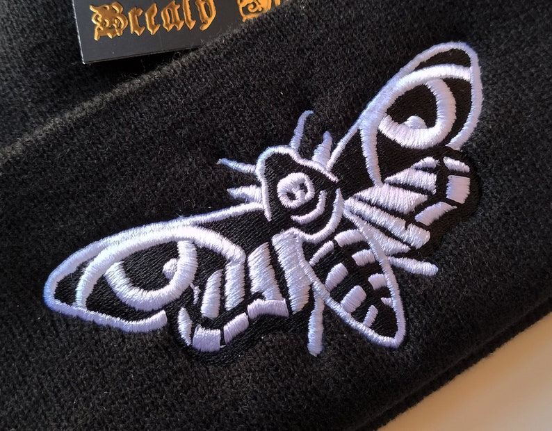 May include: Black beanie with a white embroidered moth design. The moth has a large wingspan and intricate details.