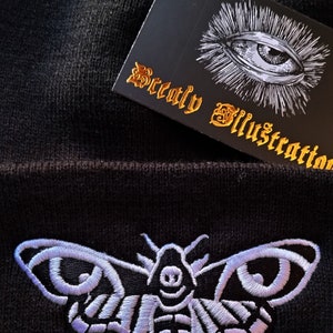 May include: Black beanie with a white embroidered death's-head hawk moth. The beanie has a tag that says "Beatly Illustration" with a white eye graphic.