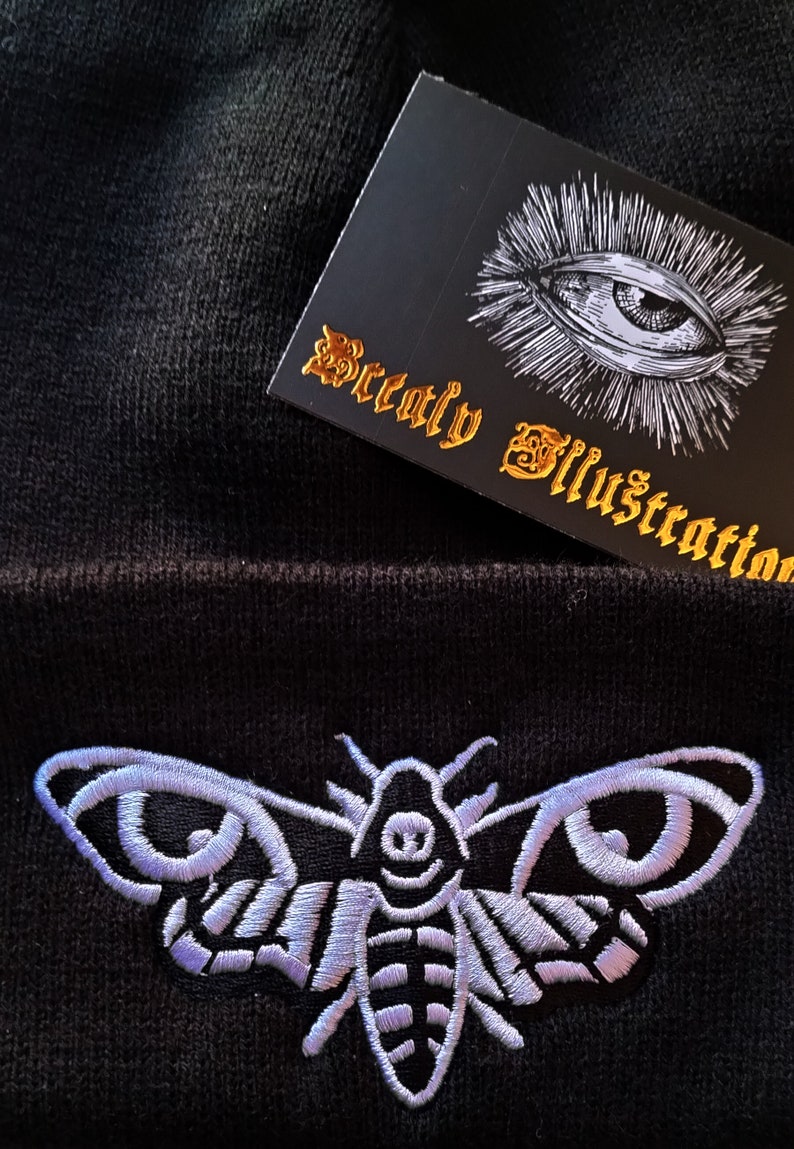 May include: Black beanie with a white embroidered death's-head hawk moth. The beanie has a tag that says "Beatly Illustration" with a white eye graphic.