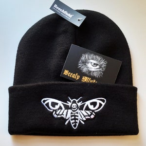May include: Black beanie with a white embroidered moth design. The beanie has a fold-over cuff and a small tag that reads "Beechfield Original Headwear".