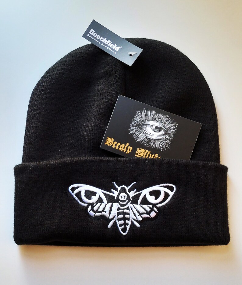 May include: Black beanie with a white embroidered moth design. The beanie has a fold-over cuff and a small tag that reads "Beechfield Original Headwear".