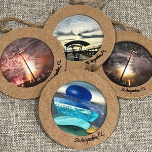 May include: Four round, brown wooden ornaments with clear glass inserts. Each ornament features a different image of St. Augustine, Florida. The images include a bridge over water, fireworks, sea glass, and a sunset.