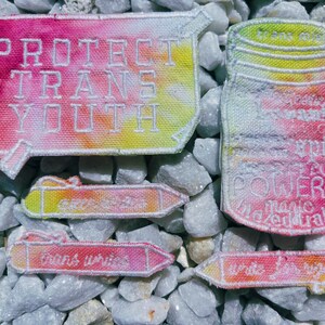 May include: A set of five embroidered iron-on patches with a tie-dye effect. The patches feature the phrases "Protect Trans Youth", "Trans Lives Matter", "Write Your Rights", "Our Power", and "Love is Magic".