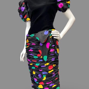May include: A black dress featuring a solid black bodice with short puffed sleeves and a ruched skirt. Both the sleeves and the skirt are decorated with a vibrant multicolored polka dot pattern in shades of purple, pink, yellow, and green. A large black fabric bow is attached at the waist.