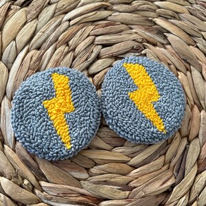 May include: Two gray and yellow round wool punch needle wall hangings with a lightning bolt design.