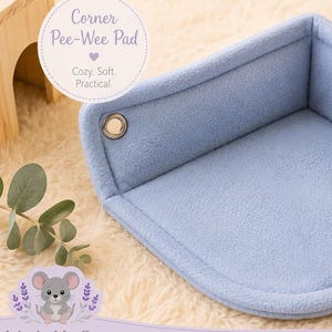 Puede incluir: Una almohadilla esquinera azul claro para mascotas pequeñas. La almohadilla está hecha de forro polar suave y tiene un diseño de esquina con dos ojales metálicos. El texto "Corner Pee-Wee Pad" está en un círculo en la esquina superior izquierda.