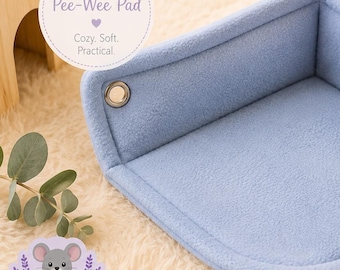 Può includere: Un cuscinetto angolare blu chiaro per piccoli animali domestici. Il cuscinetto è realizzato in morbido pile e ha un design angolare con due occhielli metallici. Il testo "Corner Pee-Wee Pad" è in un cerchio in alto a sinistra.