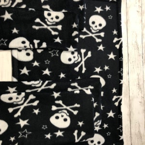 May include: Black fleece fabric with a repeating pattern of white skulls, crossbones, and stars. The fabric is folded and stacked, showing the pattern on the top and sides. The fabric is likely used for blankets or other home decor items.