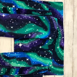 May include: A stack of soft fleece fabric pieces with a vibrant aurora borealis design. The fabric features swirling shades of blue, green, and purple, speckled with white star-like shapes. The fabric is likely used for blankets or other cozy items.