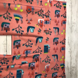 May include: A set of coral-colored fleece fabric pieces with a repeating pattern of whimsical houses, trees, and abstract shapes in shades of blue, yellow, and green. The fabric is folded and arranged to show the pattern.