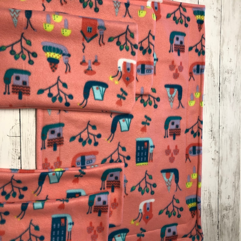 May include: A set of coral-colored fleece fabric pieces with a repeating pattern of whimsical houses, trees, and abstract shapes in shades of blue, yellow, and green. The fabric is folded and arranged to show the pattern.