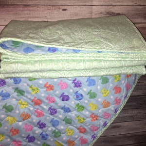 May include: A folded quilt featuring a light green top layer with a white embossed floral pattern and a light blue underside decorated with a repeating pattern of small, colorful fish in shades of yellow, pink, green, and purple on white dots.