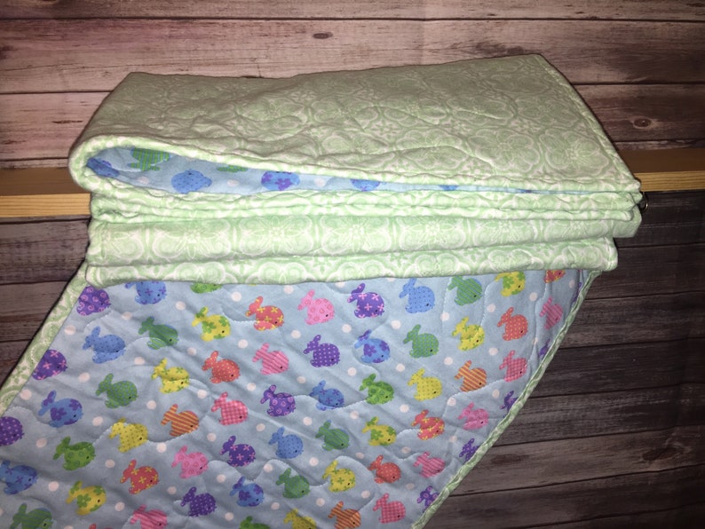 May include: A folded quilt featuring a light green top layer with a white embossed floral pattern and a light blue underside decorated with a repeating pattern of small, colorful fish in shades of yellow, pink, green, and purple on white dots.