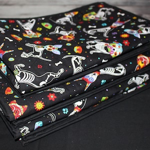 May include: A stack of folded fabric blankets featuring a black background with a repeating pattern of colorful sugar skull dog skeletons, bones, and vibrant flowers. The blankets have a solid black fabric border along the edges of the material.