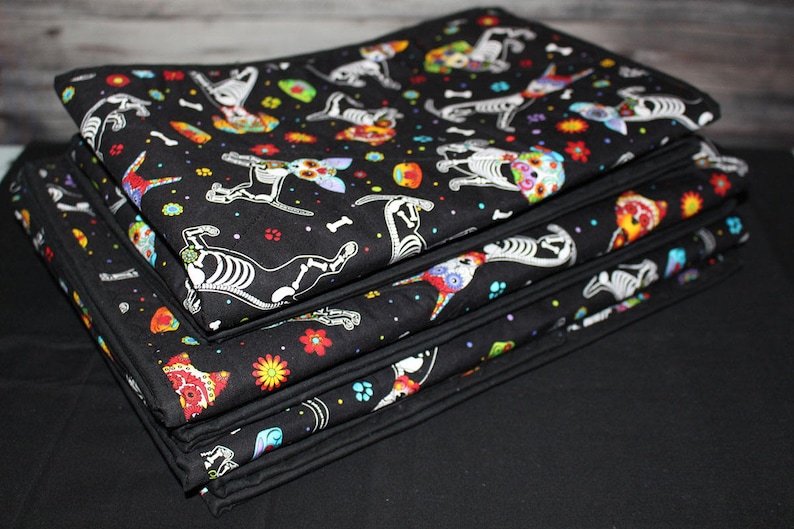 May include: A stack of folded fabric blankets featuring a black background with a repeating pattern of colorful sugar skull dog skeletons, bones, and vibrant flowers. The blankets have a solid black fabric border along the edges of the material.