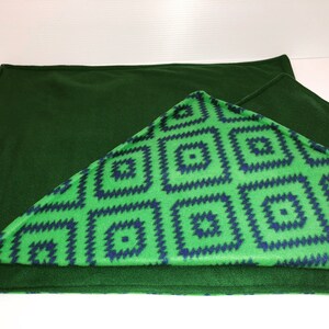 May include: A rectangular fleece blanket featuring a solid dark forest green side and a contrasting side with a bright lime green background patterned with repeating dark blue geometric square motifs arranged in a grid-like formation.