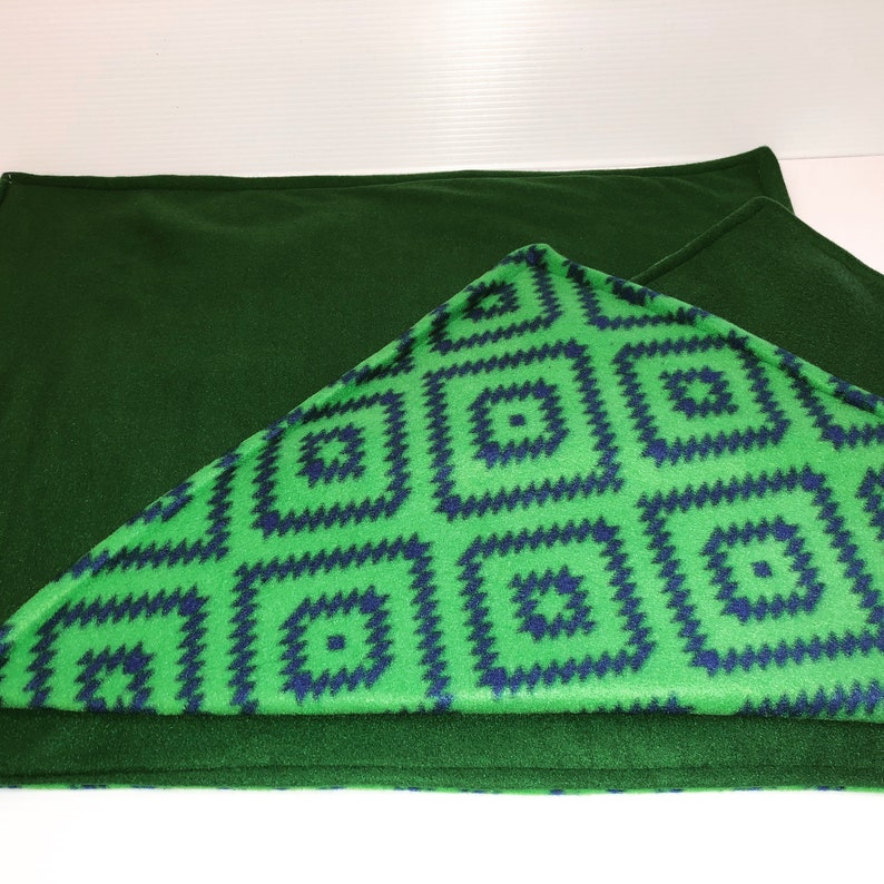 May include: A rectangular fleece blanket featuring a solid dark forest green side and a contrasting side with a bright lime green background patterned with repeating dark blue geometric square motifs arranged in a grid-like formation.