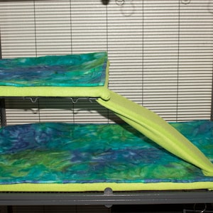 May include: A two-level pet cage liner set featuring a rectangular upper mat and a larger lower mat, both with a tie-dye pattern in shades of blue, green, and teal. A lime green ramp cover connects the two levels, providing a soft surface.