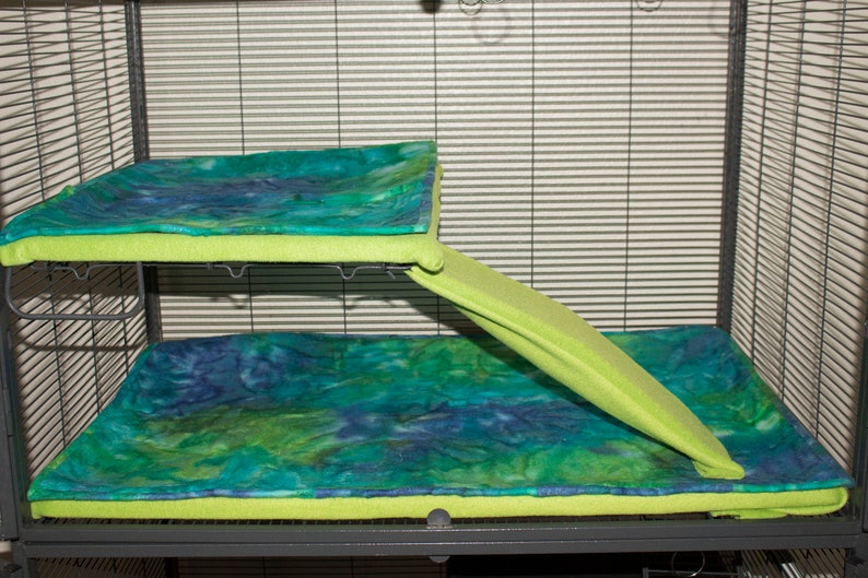 May include: A two-level pet cage liner set featuring a rectangular upper mat and a larger lower mat, both with a tie-dye pattern in shades of blue, green, and teal. A lime green ramp cover connects the two levels, providing a soft surface.