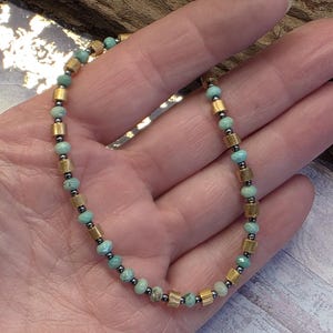 May include: A delicate bracelet featuring small, round, light turquoise beads interspersed with gold-colored cylindrical beads and tiny dark gray beads. The bracelet is displayed on a hand, showcasing its intricate design and color contrast.