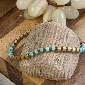May include: A beaded bracelet featuring turquoise and dark gray beads interspersed with gold-colored cylindrical beads. The bracelet is displayed on a textured, light brown stone, with a wooden background and a cluster of white, oval-shaped stones.