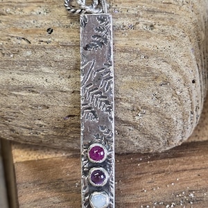 May include: A silver pendant with a rectangular design and three colorful gemstones. The pendant has a textured surface with leaf patterns. The gemstones are a deep red, purple, and light blue. The pendant is suspended from a twisted silver chain.