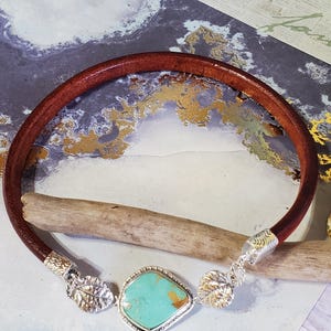 May include: A brown leather bracelet with a turquoise stone centerpiece. The stone is set in a silver-colored bezel. The bracelet has silver-colored leaf-shaped accents on each side of the stone.