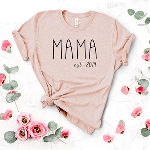 Peut inclure: Un t-shirt rose p&acirc;le avec le texte "MAMA est. 2019" en noir.