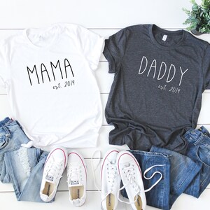 Peut inclure: Deux t-shirts, un blanc et un gris fonc&eacute;, avec les mots "Mama est. 2019" et "Daddy est. 2019" imprim&eacute;s dessus. Les t-shirts sont dispos&eacute;s sur une surface en bois blanc avec un jean bleu et des baskets blanches.