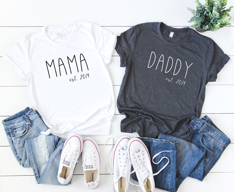 Peut inclure: Deux t-shirts, un blanc et un gris fonc&eacute;, avec les mots "Mama est. 2019" et "Daddy est. 2019" imprim&eacute;s dessus. Les t-shirts sont dispos&eacute;s sur une surface en bois blanc avec un jean bleu et des baskets blanches.