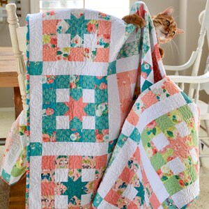 May include: A white, pink, and teal patchwork quilt with a floral pattern is draped over a white chair. A cat is peeking out from behind the quilt.