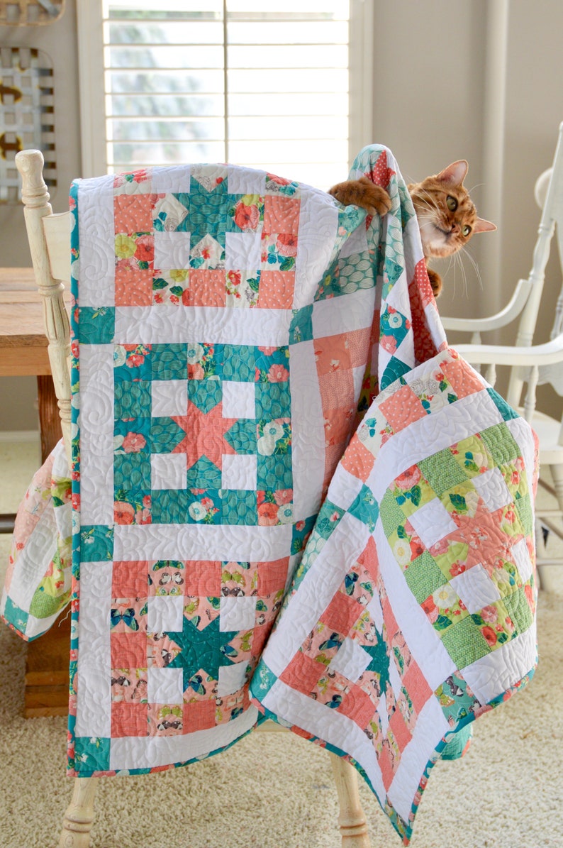 May include: A white, pink, and teal patchwork quilt with a floral pattern is draped over a white chair. A cat is peeking out from behind the quilt.