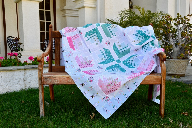 K&ouml;nnte beinhalten: Eine wei&szlig;e, rosafarbene und t&uuml;rkisfarbene Patchwork-Decke mit Blumenmuster ist gefaltet und &uuml;ber eine Holzbank gelegt. Die Bank steht in einem Garten mit gr&uuml;nem Gras und Blumen.